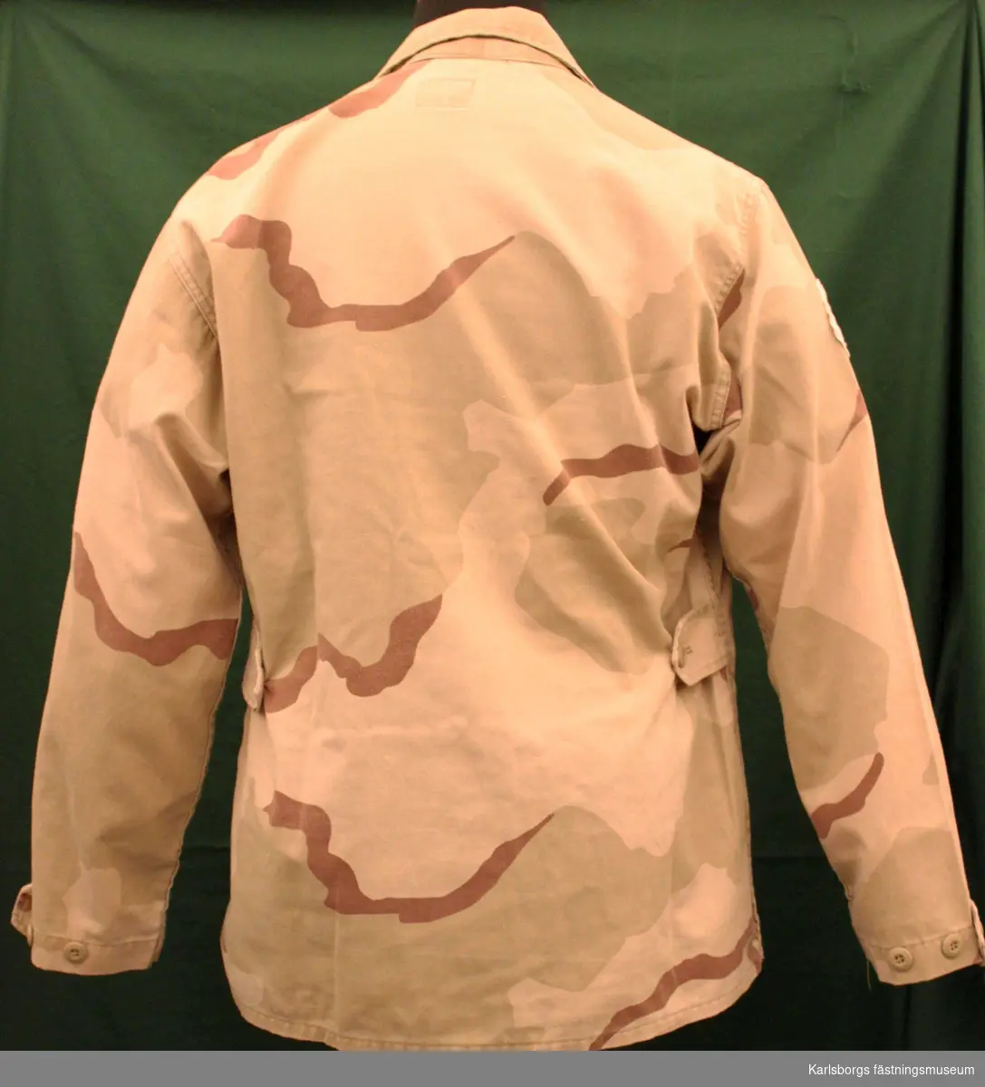 Kamouflagejacka,enkelknäppt med doldknäppning.och skjortkrage. Utanpåsydda fickor med ficklock, Röda korsemblem på höger ärm samt märke med tre kronor på vänster ärm.