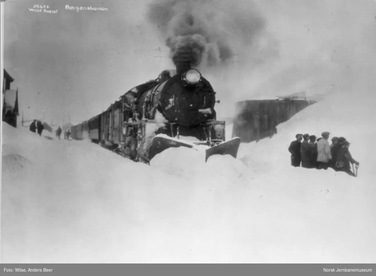 Damplokomotiv type 31b med stor frontplog foran østgående persontog på Finse stasjon : en gjeng snømåkere til høyre i bildet