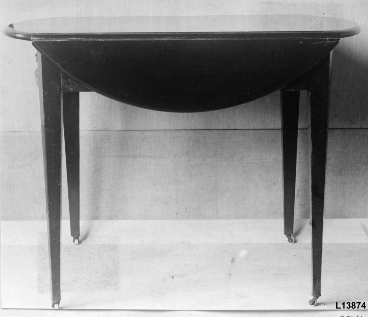 Rektangulært bord med to sideplater som kan slås opp. I oppslått posisjon er bordplaten rund. En skuff under bordplaten. Trinser under ben. 
Lysbåndintarsia langs kanten av sargen på begge kortsider.