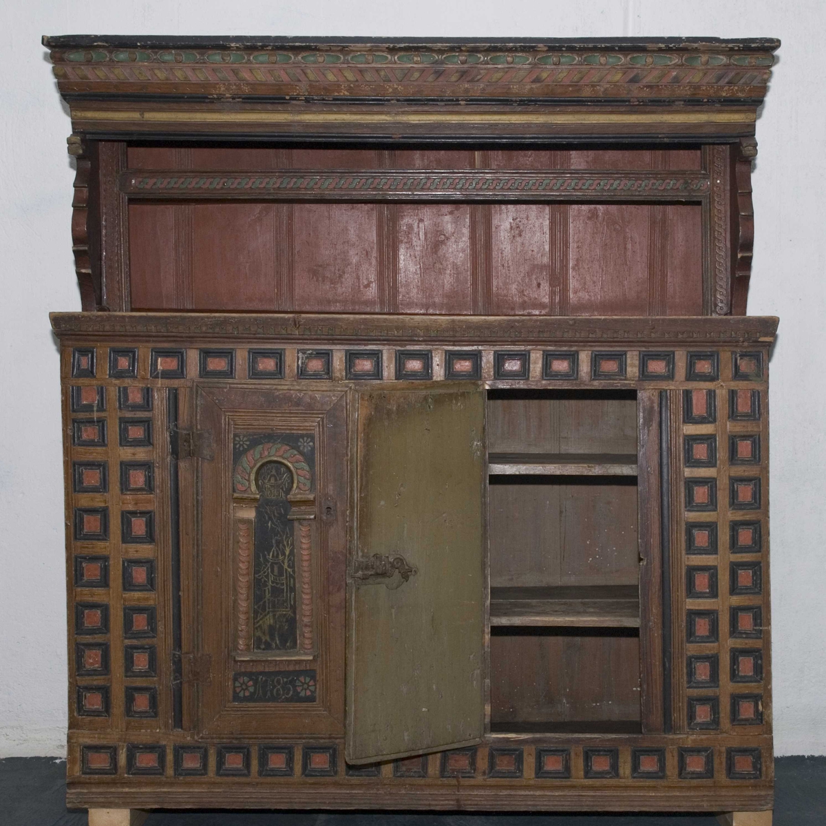 Skap i to deler. Høy skjenkebord, lav overdel. Skåret dekor i gesims med dragehoder i konsollene. Kassetter med rammefyllinger som dekor i frontpanelene. På dørene er det også malte motiv innen romanske buer. På høyre dør malte bygninger, på venstre dør er motivet uleselig. Ådret på sidene.