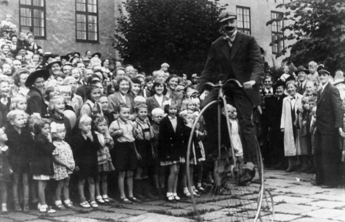 Sykkelkavalkaden på Norsk Folkemuseum i 1951. Publikum på Torvet ser på syklst på gammel sykkel.