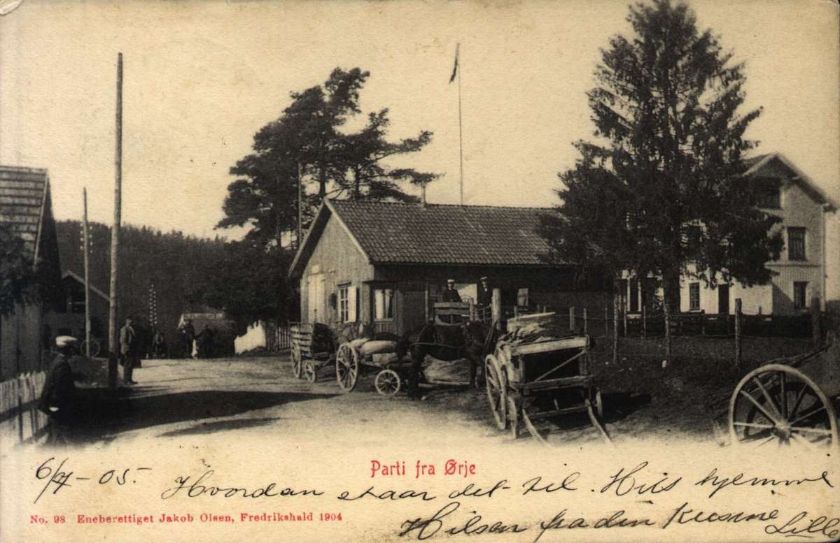 Postkort. Parti fra Ørje. Hestevogner står ved veien, flere hus ligger bak et gjerde. Stemplet 07.07.1905.