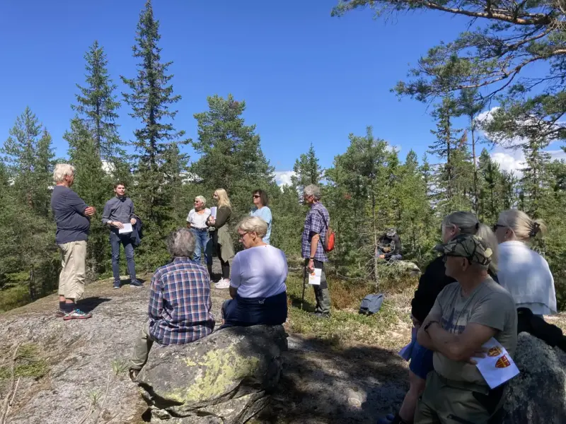 Tolv personer i skogen. De er på Olsokvandring. De hører på presten fortelle om olsoktradisjoner.