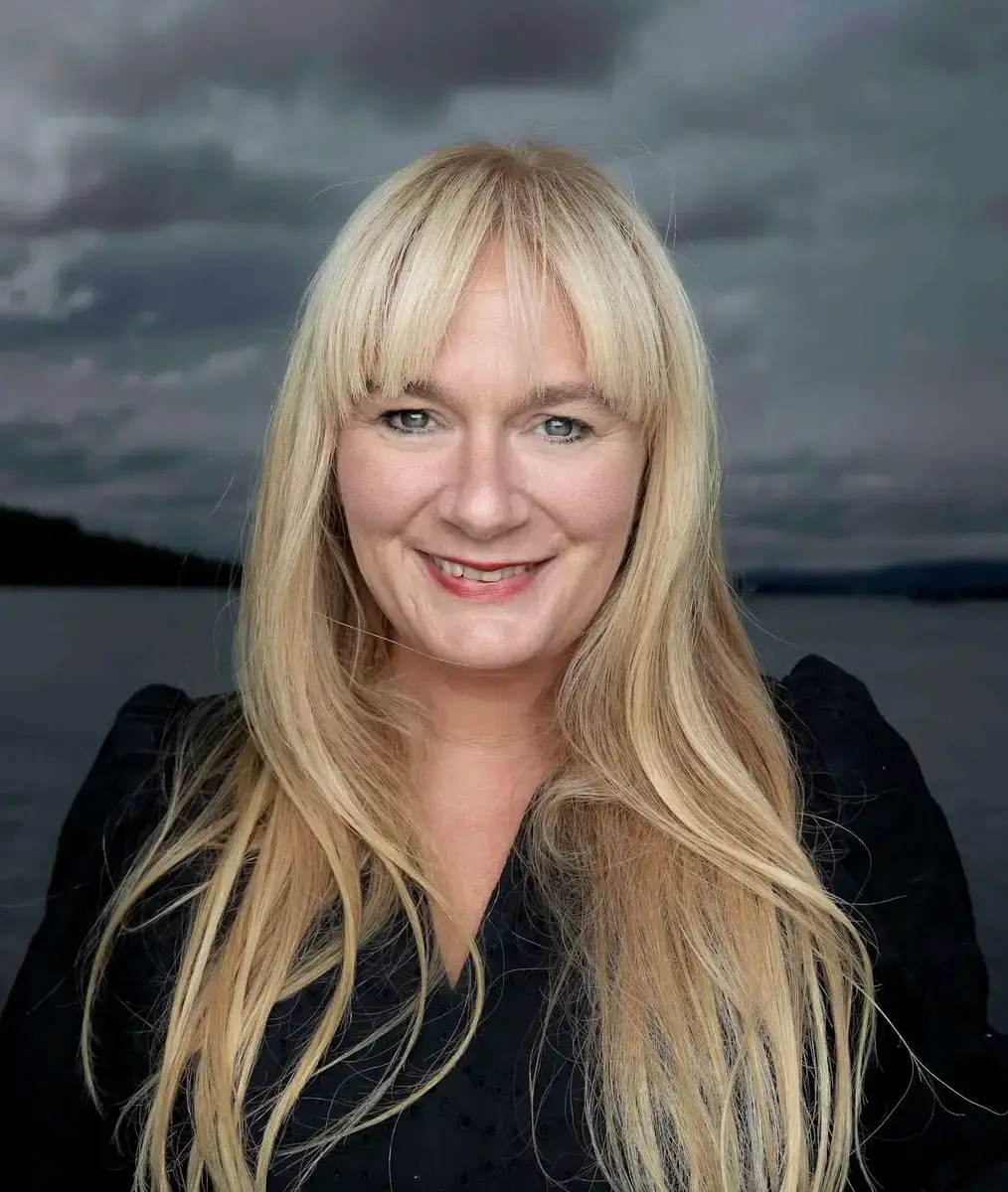 Portrett av Marit Eikemo som ser i kamera og smiler med mørk bakgrunn med sjø og himmel med skyer.