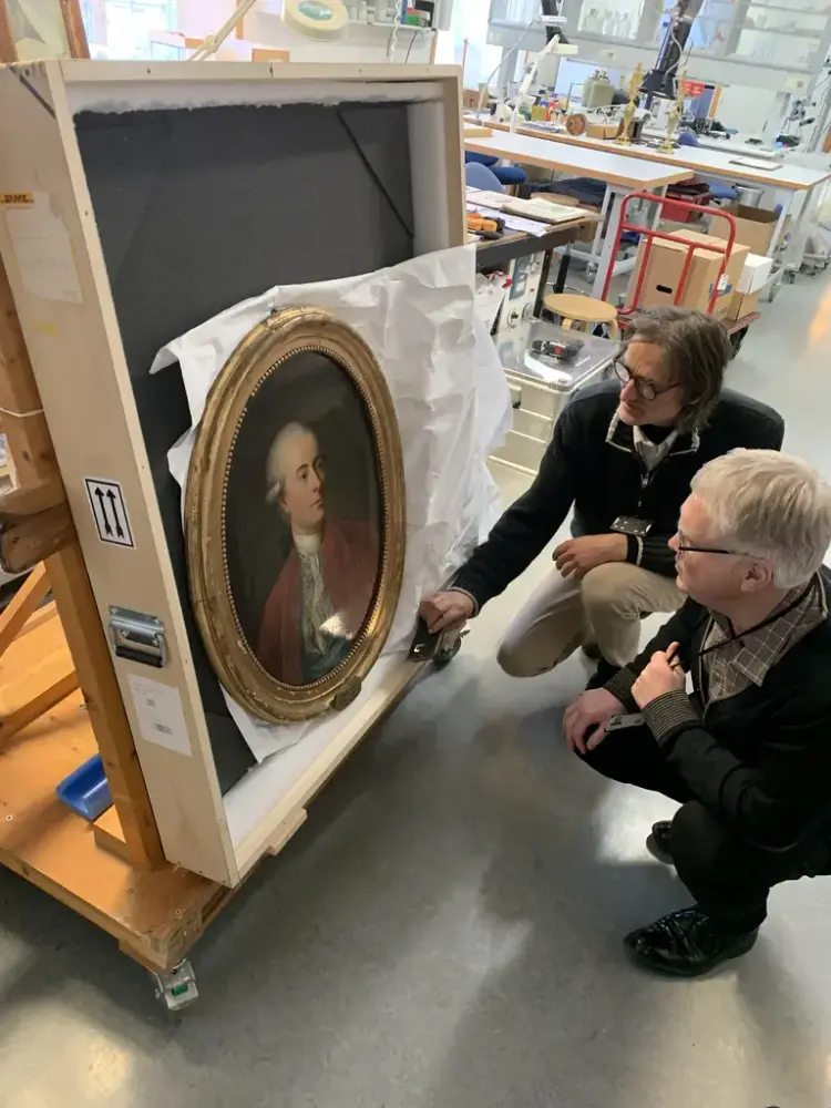 Malerikonservator Niels Gerhard Johansen og konservator Geir Thomas Risåsen sitter på huk og undersøker et ovalt portrettmaleri av Carsten Anker, som er delvis pakket ut av en transportkasse.