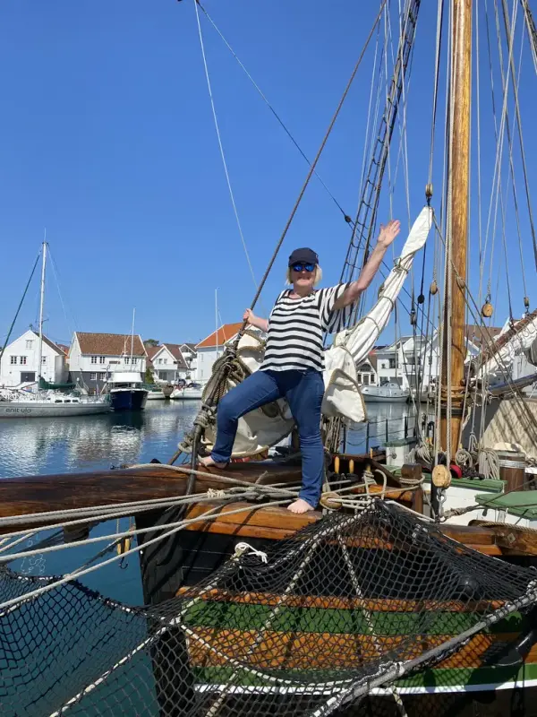 En dame i stripete skjorte står i baugen på et seilskip som ligger til kai. Hvite hus i bakgrunn.
