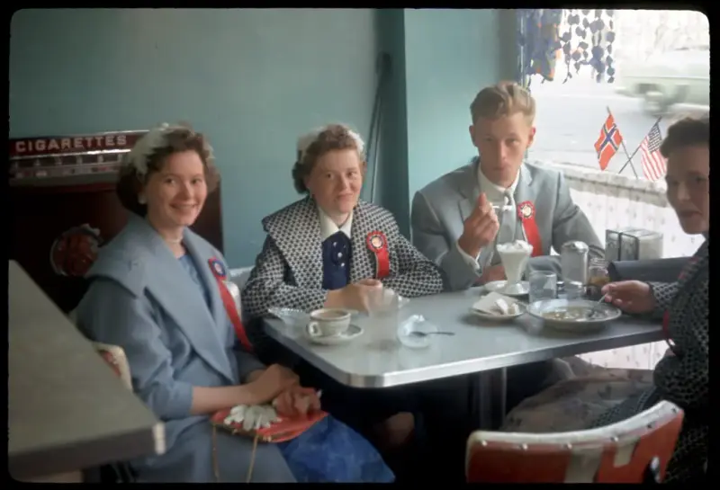 Folk på kafé på Lapskaus Boulevard, med 17. mai-sløyfer samt norsk og amerikansk flagg i vinduet.