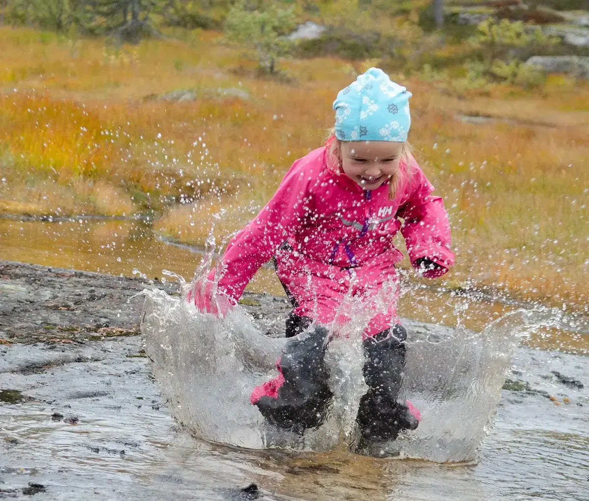 Et barn i rosa jakke og blå hatt plasker gledelig i en sølepytt, omgitt av høstlig natur. Vann buer rundt barnet mens det hopper, og fanger et øyeblikk med leken spenning.