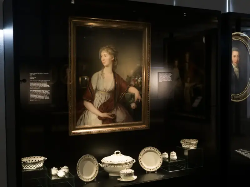 Portrait of a woman by Jens Juel, wearing a white dress with a reddish-brown shawl, holding a bouquet of flowers. Displayed in a case with white porcelain objects.